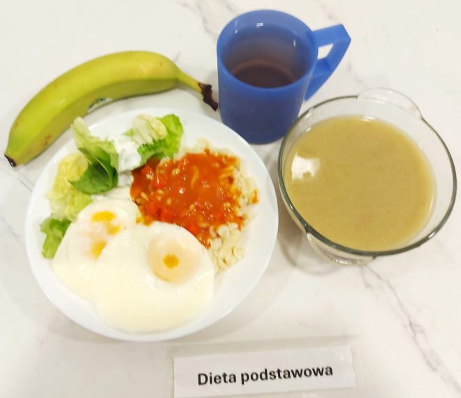 Na marmurowym blacie zaprezentowano posiłek obiadowy w ramach diety podstawowej. Na białym talerzu znajdują się dwa jajka sadzone o miękkim, lekko płynnym żółtku, porcja drobnego makaronu z jasnym sosem pomidorowym z warzywami oraz kilka liści sałaty z dodatkiem śmietany. Obok leży cały, dojrzały banan. Po prawej stronie widoczna jest miska z jasnobrązową, kremową zupą – najprawdopodobniej warzywnym kremem lub zupą z brokułów. Do obiadu podano ciemny napar (np. herbatę) w niebieskim, plastikowym kubku z uchem. Całość oznaczona etykietą „Dieta podstawowa”