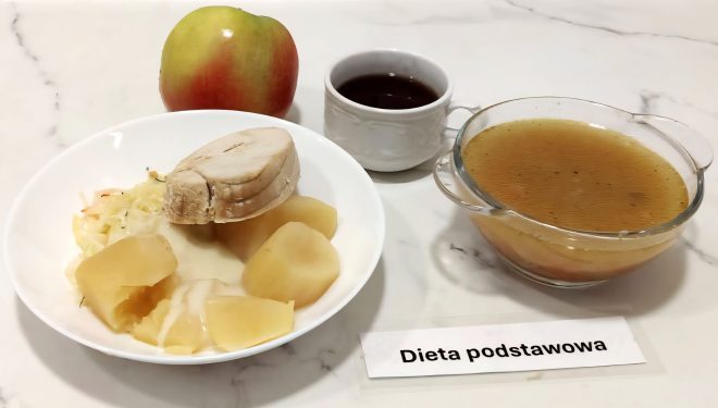 Na białym talerzu podano porcję gotowanych ziemniaków z jasnym sosem, kawałek gotowanego mięsa drobiowego oraz surówkę z białej kapusty. Obok talerza stoi filiżanka z ciemnym kompotem, świeże jabłko oraz szklana miseczka z porcją rosołu z drobnymi cząstkami warzyw. Całość ułożona na marmurowym blacie. Przy posiłku znajduje się podpis „Dieta podstawowa”