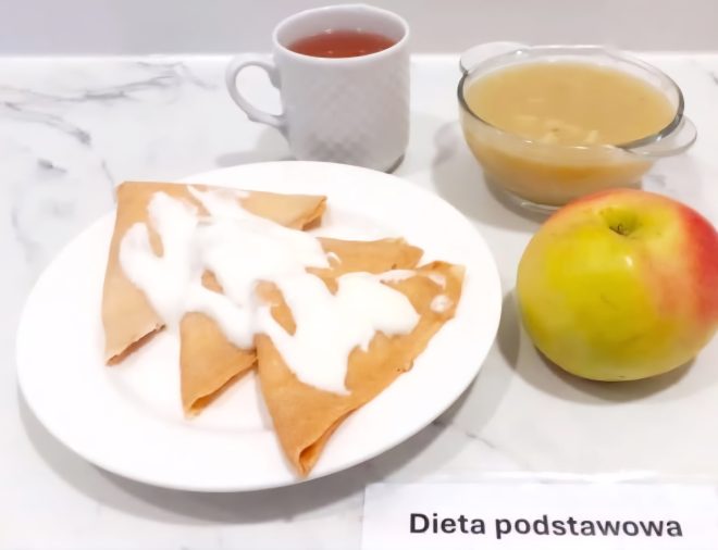 Posiłek obiadowy serwowany w diecie podstawowej. Na talerzu trzy naleśniki złożone w trójkąty, polane białym sosem jogurtowym lub śmietanowym. Obok filiżanka z czerwonym naparem, szklana salaterka z zupą jarzynową oraz żółtoczerwone jabłko. Na blacie po prawej stronie kartka z podpisem „Dieta podstawowa”.