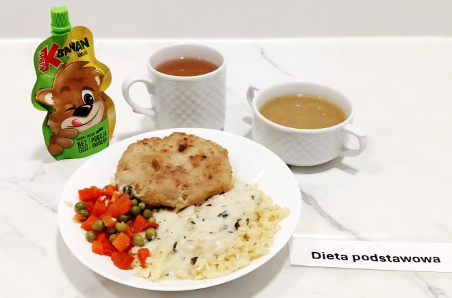 Obiad serwowany na białym talerzu: panierowany kotlet, gotowana marchewka z groszkiem oraz drobny makaron polany jasnym sosem koperkowym. W tle dwa białe kubki – jeden z herbatą, drugi z jasnym bulionem. Obok opakowanie przecieru owocowego dla dzieci z nadrukiem uśmiechniętego misia i napisem „Banan Jabłko”. Na blacie znajduje się też etykieta z napisem: „Dieta podstawowa”
