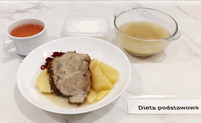 Zestaw obiadowy w diecie podstawowej serwowany na białym talerzu. Na danie główne składają się gotowane ziemniaki, plaster pieczeni wieprzowej w jasnym sosie oraz duszona czerwona kapusta. W tle znajduje się bulion w szklanym naczyniu, kompot w białym kubku oraz deser mleczny w plastikowym pojemniku. Całość umieszczona na marmurowym blacie, obok znajduje się etykieta z napisem „Dieta podstawowa”