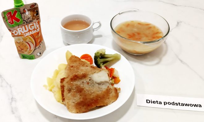 Talerz z obiadem w diecie podstawowej – panierowany filet rybny, gotowane ziemniaki oraz mieszanka warzyw (brokuł, marchew, kalafior). Obok miseczka z zupą warzywną, kubek z kompotem oraz mus owocowy „Kubuś” w saszetce. Całość ustawiona na białym stole, z karteczką "Dieta podstawowa"