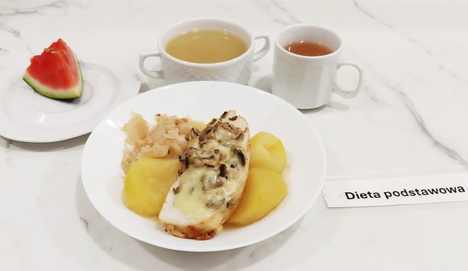 Obiad w diecie podstawowej: filet z kurczaka zapiekany z pieczarkami i serem, gotowane ziemniaki oraz kapusta kiszona. Obok kawałek arbuza na osobnym talerzyku. Do picia: filiżanka herbaty oraz bulion w białym kubeczku z uszkiem