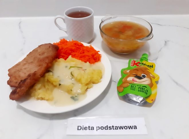 Talerz z panierowanym kotletem, ziemniakami z sosem koperkowym i tartą marchewką. Obok miseczka zupy jarzynowej, filiżanka herbaty i mus owocowy w tubce z rysunkiem misia. Dieta podstawowa