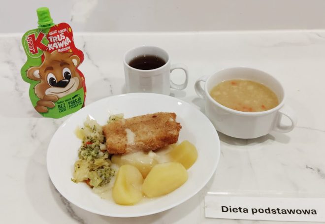 Obiad w diecie podstawowej: na głównym talerzu znajdują się trzy ziemniaki, panierowany filet rybny z sosem oraz mieszanka gotowanych warzyw (m.in. brokuły, kapusta, marchew). Towarzyszy mu zupa jarzynowa w białej miseczce oraz kubek z herbatą. W tle znajduje się mus owocowo-warzywny w saszetce dla dzieci o smaku truskawkowo-marchewkowym. Całość podana na jasnym blacie, z etykietą „Dieta podstawowa”