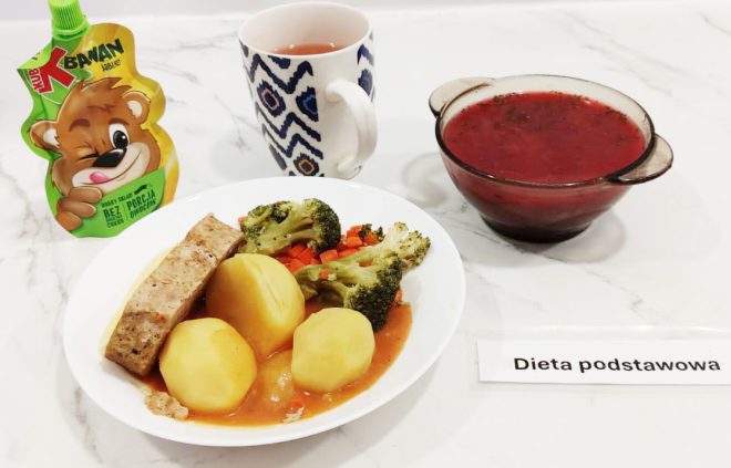Obiad w diecie podstawowej: na talerzu trzy ziemniaki, plaster pieczeni, gotowane brokuły i marchewka w sosie. Obok kubek z herbatą, saszetka przecieru jabłkowego oraz miseczka z czerwonym barszczem. Napis: Dieta podstawowa