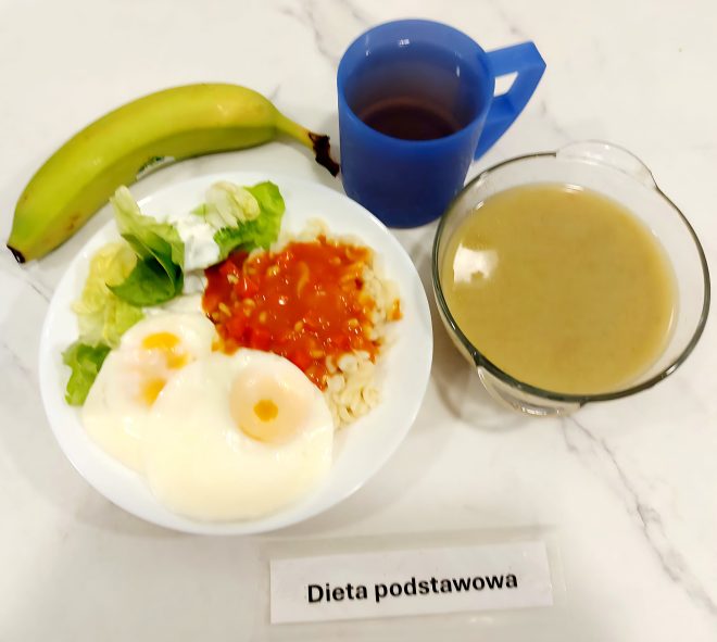Na zdjęciu znajduje się zestaw obiadowy serwowany w ramach diety podstawowej. Na talerzu podano dwa sadzone jajka na miękko, porcję drobnego makaronu z sosem pomidorowym oraz dodatek świeżej sałaty z odrobiną śmietany. Obok leży cały banan. Po prawej stronie znajduje się miska z zupą kremem – prawdopodobnie z warzyw korzeniowych lub kalafiora – o gładkiej konsystencji, bez widocznych kawałków składników. Do posiłku podano także ciemny napój w niebieskim plastikowym kubku z uchem, najpewniej herbatę owocową. Całość umieszczona jest na białym blacie z etykietą „Dieta podstawowa”