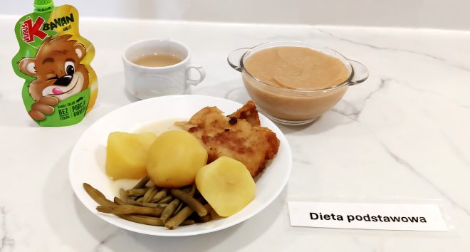 Na marmurowym blacie ustawiono zestaw obiadowy dla dzieci lub pacjentów objętych dietą podstawową. Na białym talerzu znajduje się porcja gotowanych ziemniaków, panierowany filet rybny oraz zielona fasolka szparagowa. Obok ustawiono szklane naczynie z musem jabłkowym lub owocowym oraz filiżankę z jasnym napojem, prawdopodobnie kawą zbożową lub herbatą z mlekiem. Dodatkowym elementem jest owocowy mus jabłkowy w tubce z wizerunkiem misia (marki Kubuś). Zestaw oznaczony etykietą „Dieta podstawowa”