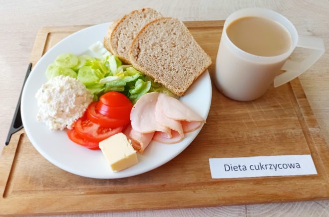 Na białym talerzu podanym na drewnianej tacy znajduje się śniadanie w wersji diety cukrzycowej. W skład posiłku wchodzą: trzy plasterki pieczywa razowego, masło, plastry chudej wędliny z kurczaka lub indyka, biały ser ziarnisty (twarożek), plasterki ogórka, liście sałaty oraz plastry pomidora. Obok talerza stoi kubek z napojem, najprawdopodobniej kawa z mlekiem. Na tacy umieszczona etykieta z napisem „Dieta cukrzycowa”