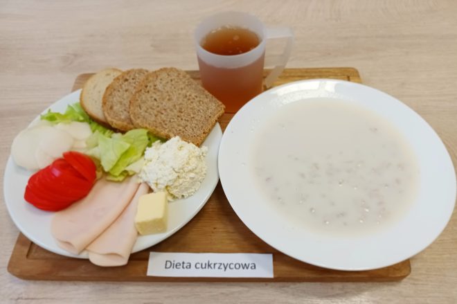 Taca z zestawem śniadaniowym podpisanym jako "Dieta cukrzycowa", ułożona na jasnym stole. Na pierwszym planie biały talerz z porcją białego sera, dwiema plastrami szynki drobiowej, kostką masła, liśćmi sałaty, plastrami pomidora i rzodkiewki oraz trzema kromkami pieczywa pełnoziarnistego. Obok głębokiego talerza znajduje się jasna zupa mleczna z drobnymi płatkami, najprawdopodobniej zbożowymi. Z tyłu, pośrodku tacy, plastikowy przezroczysty kubek z jasnobrązowym napojem – prawdopodobnie herbatą. Całość estetycznie podana, z dbałością o zbilansowanie składników odpowiednich dla osób z cukrzycą