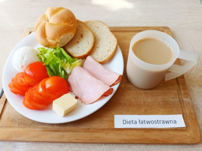 Na zdjęciu ukazano śniadanie w ramach „Diety łatwostrawnej” ułożone na jasnej tacy. Biały talerz zawiera bułkę pszenną typu kajzerka, dwie kromki jasnego pieczywa, plasterki szynki drobiowej, kilka plastrów pomidora, kulkę białego twarogu, listek sałaty oraz niewielką kostkę masła. Towarzyszy im napój przypominający kawę z mlekiem, podany w przezroczystym kubku z uchwytem. Estetyczne i lekkie podanie sugeruje posiłek odpowiedni dla osób z dolegliwościami żołądkowo-jelitowymi