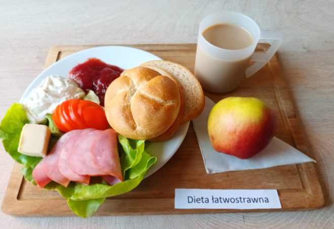 Taca ze śniadaniem w ramach diety łatwostrawnej. Na talerzu znajduje się bułka kajzerka, kromka białego pieczywa, plasterki szynki, liść sałaty, plastry pomidora, porcja twarożku i masła oraz galaretka z czerwonych owoców. Obok na tacy: kubek z kawą z mlekiem oraz czerwono-żółte jabłko na serwetce. Na desce widoczna etykieta „Dieta łatwostrawna”