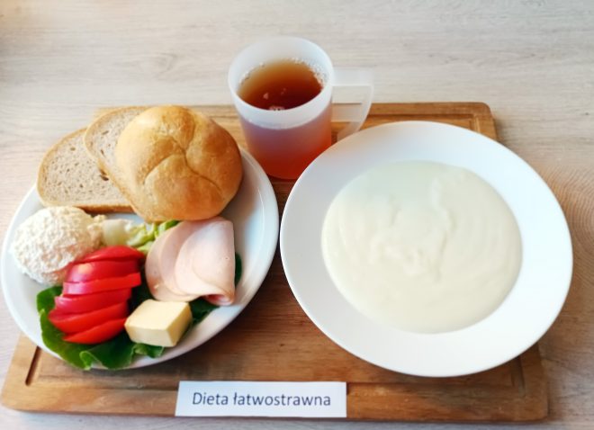 Śniadanie serwowane w ramach diety łatwostrawnej na drewnianej tacy, z widoczną kartką z napisem „Dieta łatwostrawna”. Po lewej stronie talerz z pieczywem: jasną bułką typu kajzerka oraz dwoma kromkami chleba pszennego. Obok plasterki chudej wędliny drobiowej, porcja twarogu, plasterek żółtego sera, kilka kawałków pomidora i liść sałaty. Po prawej stronie głęboki talerz z zupą mleczną o jednolitej, gładkiej konsystencji. W górnej części tacy plastikowy kubek z jasną herbatą.