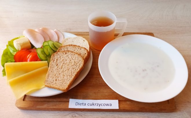Taca z podpisem „Dieta cukrzycowa”. Na niej śniadanie składające się z białej miski z mleczną zupą z drobną kaszą, kubka z herbatą lub naparem ziołowym, oraz talerza z trzema kromkami pieczywa (ciemnego i jasnego), plasterkami wędliny z indyka, żółtym serem, sałatą, pomidorem, ogórkiem i porcją masła