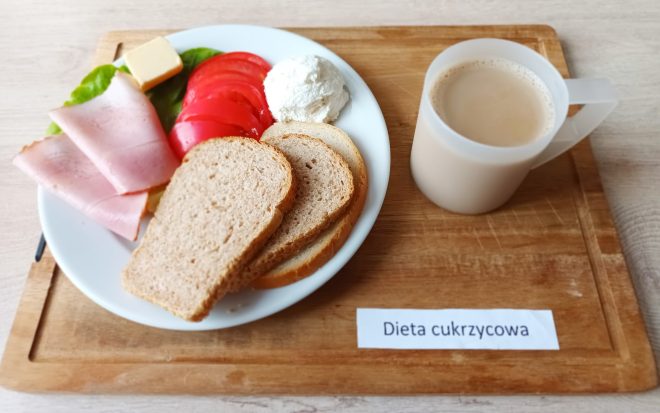Śniadanie w diecie cukrzycowej: talerz z trzema kromkami pieczywa mieszanego, plasterkami szynki, pomidora, kawałkiem masła i porcją białego twarożku, podanymi na sałacie. Obok kubek z kawą z mlekiem lub napojem mlecznym. Na tacy znajduje się etykieta z napisem „Dieta cukrzycowa”