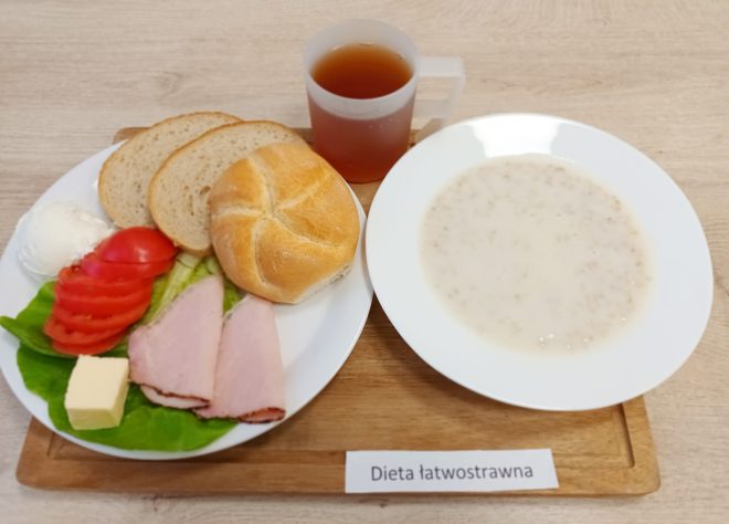 Taca z posiłkiem oznaczonym jako „Dieta łatwostrawna”. Na niej: talerz z białym pieczywem (kajzerka i dwie kromki chleba), dwie plasterki szynki, plasterki pomidora, sałata, masło i twarożek. Obok miska z mleczną zupą (prawdopodobnie owsianką) oraz kubek z herbatą. Wszystko podane na drewnianej podstawce