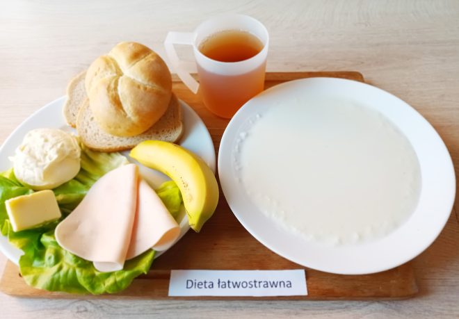 Taca z posiłkiem śniadaniowym oznaczonym jako „Dieta łatwostrawna”. Na talerzu bułka kajzerka i dwie kromki białego pieczywa, plaster szynki drobiowej, banan, masło, porcja białego twarożku na sałacie. Obok znajduje się kubek z jasną herbatą oraz talerz z gładką białą kaszką mleczną lub kleikiem ryżowym