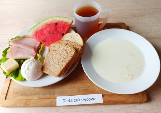 Zestaw śniadaniowy w diecie cukrzycowej na białym talerzu i drewnianej tacy. W skład posiłku wchodzą dwa plasterki szynki, porcja białego twarogu, plaster masła, dwa kawałki chleba razowego, kromka chleba pszennego i trójkąt arbuza – wszystko ułożone na liściu sałaty. Obok miska z mlekiem i płatkami, a do picia herbata w półprzezroczystym kubku z uchem. Na tacy etykieta z napisem „Dieta cukrzycowa”