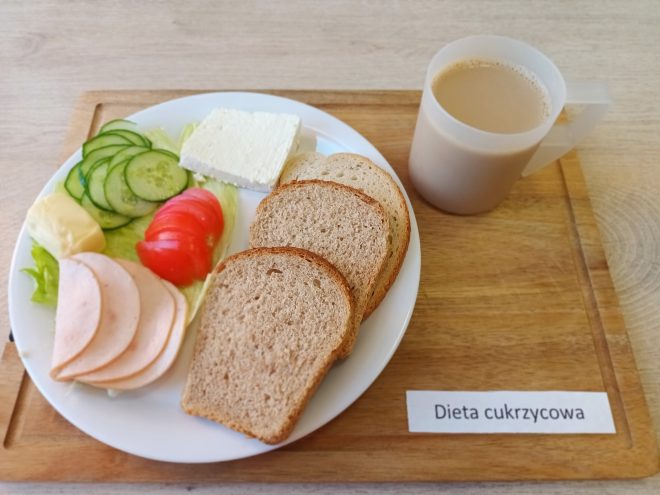 Zestaw śniadaniowy przeznaczony dla osób na diecie łatwostrawnej. Na talerzu znajduje się jasna bułka kajzerka, dwa plasterki chleba pszennego, plasterki chudej wędliny z indyka, plasterek żółtego sera, porcje twarożku i galaretki z czerwonych owoców, kilka plasterków pomidora, całe żółte jabłko oraz liście sałaty. Zupa mleczna z drobną kaszą podana jest w głębokim talerzu. Do picia – herbata w półprzezroczystym kubku z uchem. Całość ustawiona na drewnianej tacy oznaczonej etykietą „Dieta łatwostrawna”