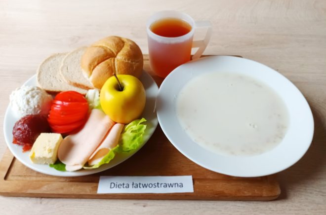 Zestaw śniadaniowy dostosowany do potrzeb osób z cukrzycą. Na talerzu znajdują się trzy kromki pieczywa razowego, plasterki wędliny drobiowej, biały ser typu feta, masło, pokrojone plasterki ogórka i pomidora oraz liście sałaty. Obok znajduje się napój mleczny lub kawa z mlekiem, podany w półprzezroczystym kubku z uchem. Całość ułożona na drewnianej podstawce oznaczonej etykietą „Dieta cukrzycowa”