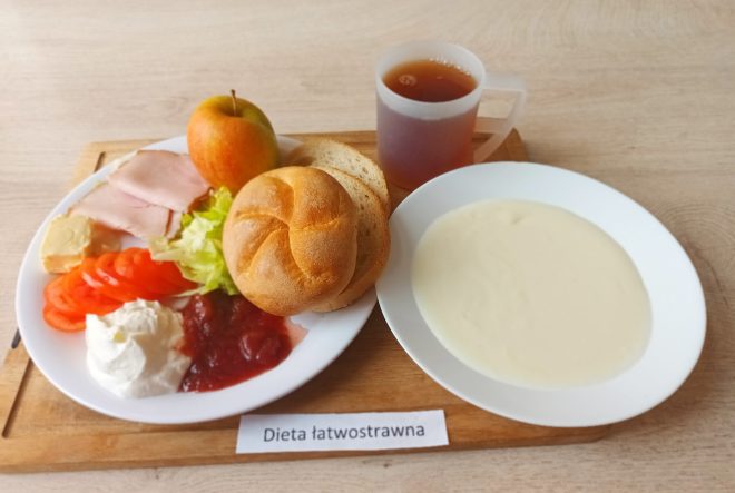 Taca ze śniadaniem w diecie łatwostrawnej: talerz z pieczywem (bułka i kromki chleba pszennego), plastrem szynki drobiowej, masłem, sałatą, pomidorem, twarożkiem, musem owocowym oraz całym jabłkiem. Obok talerz z kremową zupą (prawdopodobnie zupa mleczna lub z kaszą manną) i kubek z herbatą. Na tacy widoczna etykieta „Dieta łatwostrawna”