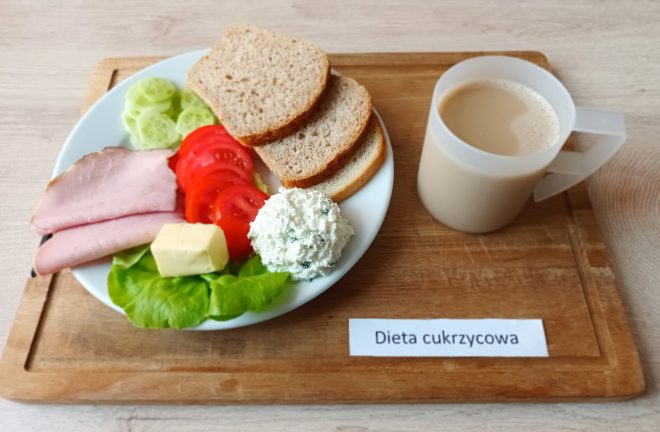 Śniadanie w diecie cukrzycowej: talerz z trzema kromkami chleba razowego, plastrem szynki, plasterkami ogórka i pomidora, kostką masła, porcją twarożku z ziołami oraz liściem sałaty. Obok kubek z kawą z mlekiem. Całość ustawiona na drewnianej tacy z podpisem „Dieta cukrzycowa”
