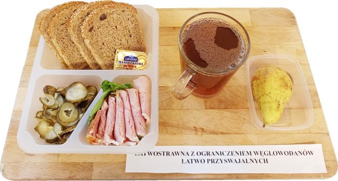 Zestaw śniadaniowy na drewnianej tacy, oznaczony jako „Dieta łatwostrawna z ograniczeniem węglowodanów łatwo przyswajalnych”. W plastikowym pojemniku z trzema przegrodami znajdują się: kilka kromek pieczywa wieloziarnistego z ziarnami sezamu i lnu, opakowanie masła, plasterki szynki z kurczaka ułożone na liściu sałaty oraz porcja ogórków konserwowych w plasterkach. Obok pojemnika stoi szklanka ciemnego naparu – najpewniej herbaty – oraz plastikowy pojemnik z połówką gruszki. Kompozycja lekkostrawna, z niskim indeksem glikemicznym