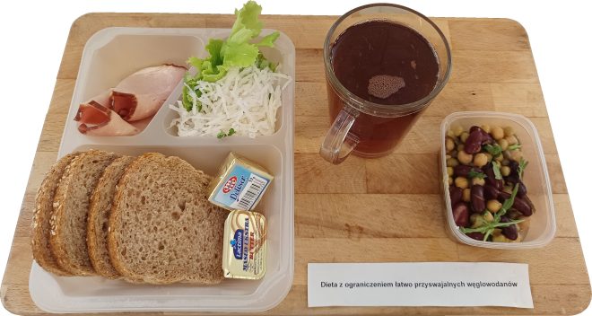 Śniadanie oznaczone jako „Dieta z ograniczeniem łatwo przyswajalnych węglowodanów”. Na tacy znajduje się kilka kromek chleba pełnoziarnistego z ziarnami, dwa plasterki wędlin (szynka i dojrzewająca), porcja białej surówki (prawdopodobnie z selera i sałaty), dwa opakowania masła, przezroczysty kubek z herbatą oraz oddzielny pojemnik z sałatką z trzech rodzajów fasoli i cieciorki z dodatkiem natki pietruszki. Na desce widoczna jest etykieta z nazwą diety