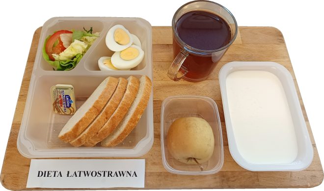 Śniadanie w ramach diety łatwostrawnej ułożone na drewnianej tacy. W plastikowym pojemniku: trzy kromki białego pieczywa, plasterki gotowanego jajka, liście sałaty z plastrem pomidora i ogórka, kostka masła. Obok: szklanka czarnej herbaty, pojemniczek z jasnym jabłkiem oraz oddzielny pojemnik z białym produktem mlecznym – najprawdopodobniej jogurt naturalny lub kefir. Na tacy widoczna etykieta „DIETA ŁATWOSTRAWNA”.
