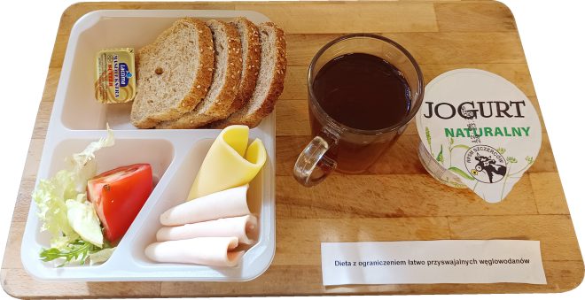 Taca z podpisem „Dieta z ograniczeniem łatwo przyswajalnych węglowodanów”. Na niej plastikowa tacka z trzema kromkami pieczywa razowego z ziarnami, porcją masła, plasterkami wędliny z indyka, żółtym serem, sałatą i kawałkiem pomidora. Obok szklanka z herbatą bez cukru oraz kubek jogurtu naturalnego