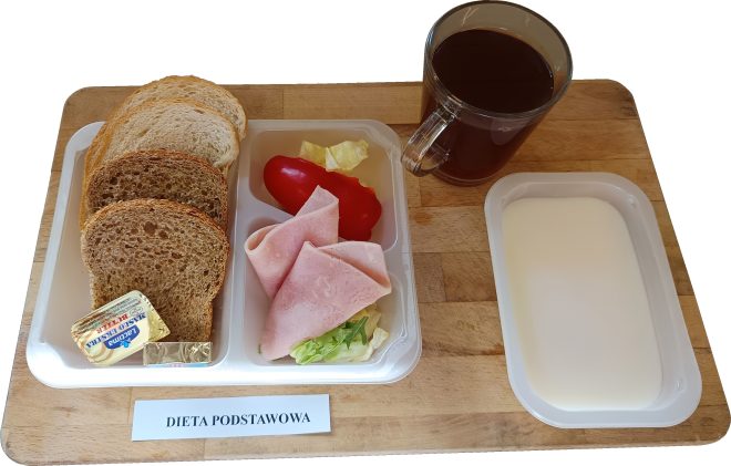 Standardowe śniadanie w ramach diety podstawowej, skierowane do pacjentów bez szczególnych ograniczeń żywieniowych. Zestaw łączy w sobie produkty zbożowe, białkowe i warzywa. Skład zestawu: Pieczywo: Trzy kromki – jedna jasna pszenna i dwie pełnoziarniste (razowe), dostarczające energii i błonnika. Dodatek białkowy: Plasterki chudej wędliny drobiowej (najpewniej z szynki z indyka) podane na sałacie. Warzywa: Kawałek czerwonej papryki oraz liście sałaty. Tłuszcz: Porcja masła "Łaciate" – jako źródło tłuszczu i witamin rozpuszczalnych w tłuszczach. Napój: Kubek kawy zbożowej lub herbaty bez cukru. Dodatkowy składnik: Pojemnik z białym napojem – prawdopodobnie mleko lub napój mleczny naturalny, jako źródło wapnia. Charakterystyka diety: Posiłek urozmaicony i zbilansowany. Zawiera produkty z każdej grupy żywieniowej. Brak specjalnych ograniczeń – odpowiedni dla ogólnej populacji pacjentów