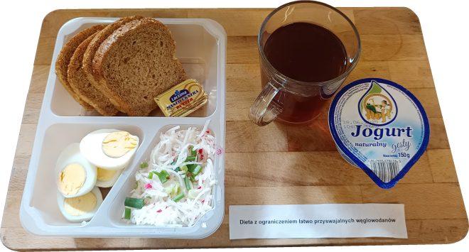 Taca z dietą o ograniczonej zawartości łatwo przyswajalnych węglowodanów. Na niej: trzy kromki chleba pełnoziarnistego, porcja masła w kostce (Lactima), dwa jajka na twardo pokrojone w plasterki, surówka z białej rzodkwi z dodatkiem szczypiorku i rzodkiewki, kubek herbaty bez cukru oraz opakowanie jogurtu naturalnego gęstego marki „Hej”. Na tacy widoczna też karteczka z napisem: „Dieta z ograniczeniem łatwo przyswajalnych węglowodanów”