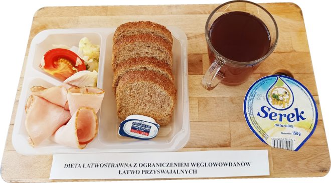 Taca ze śniadaniem w diecie łatwostrawnej z ograniczeniem węglowodanów łatwo przyswajalnych. Na tacy znajduje się pojemnik z pieczywem pełnoziarnistym, plasterkami szynki drobiowej, sałatką z pomidorem i kapustą oraz porcją masła. Obok znajduje się naturalny serek homogenizowany i szklanka czerwonej herbaty. U dołu widoczna etykieta z nazwą diety