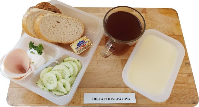 Taca ze śniadaniem szpitalnym zgodnym z dietą podstawową. Zestaw obejmuje: Trzy kromki pieczywa pszennego i żytniego, Porcję masła w małym opakowaniu, Plasterek chudej wędliny drobiowej, Porcję białego twarożku ze szczypiorkiem i natką pietruszki, Pokrojone plasterki świeżego ogórka z ziołami, Jasny napój mleczny (prawdopodobnie mleko lub kisiel mleczny), Kubek z herbatą bez dodatków. Posiłek podany w plastikowych przegródkowych pojemnikach na desce z kartką z napisem „Dieta podstawowa”