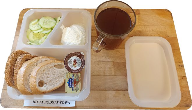 Śniadanie podane w pojemniku: plasterki jasnego i ciemnego pieczywa, biały ser twarogowy, plasterki ogórka, masło i porcja kremu czekoladowo-orzechowego. Obok szklanka kawy zbożowej i miseczka mleka. Etykieta: „DIETA PODSTAWOWA”