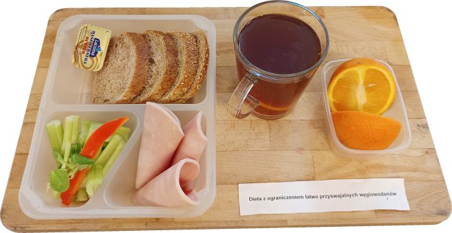 Zestaw śniadaniowy dostosowany do diety z ograniczoną ilością cukrów prostych. Na podzielonym plastikowym talerzu znajdują się trzy kromki pełnoziarnistego pieczywa z ziarnami, porcja margaryny w jednorazowym opakowaniu, plasterki szynki drobiowej, świeże warzywa – łodygi selera naciowego i pasek czerwonej papryki – oraz liście roszponki. W osobnym pojemniczku – dwa plasterki pomarańczy. Obok postawiono szklankę z gorącym naparem (herbata lub napój ziołowy). Całość znajduje się na drewnianej tacy z kartką podpisaną: „Dieta z ograniczeniem łatwo przyswajalnych węglowodanów”