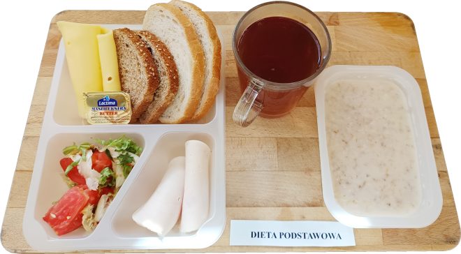 aca z przegródkami zawierająca trzy kromki pieczywa mieszanego, dwie rolki żółtego sera, plasterki szynki drobiowej, porcję masła w opakowaniu, sałatkę z pomidora i sałaty. Obok znajduje się przezroczysty kubek z czerwonym naparem i oddzielny pojemnik z owsianką na mleku. Etykieta na tacy: "Dieta podstawowa"