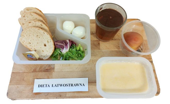 Śniadanie w diecie łatwostrawnej podane w pojemnikach: pieczywo pszenne, masło, dwa jajka na twardo oraz miks sałat z plasterkiem szynki. Obok w osobnych naczyniach: grysik na mleku, herbata w szklance oraz zapakowana gruszka. Posiłek ustawiony na tacy z podpisem „DIETA ŁATWOSTRAWNA”