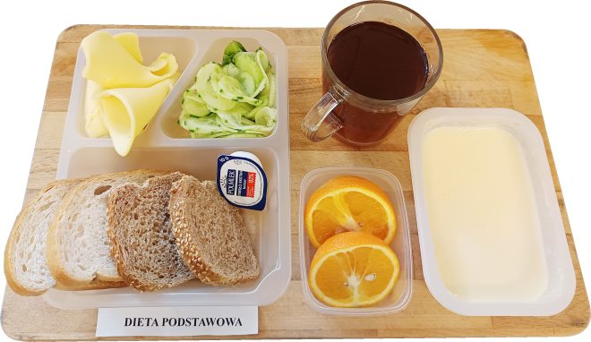 Zestaw śniadaniowy na tacy w diecie podstawowej: trzy kromki chleba pszennego i razowego, plasterki żółtego sera, sałatka z ogórka, porcja masła, dwa kawałki pomarańczy, kubek z gorącą herbatą oraz pojemniczek z białym jogurtem lub mlekiem zsiadłym. Widoczna etykieta z napisem „DIETA PODSTAWOWA”