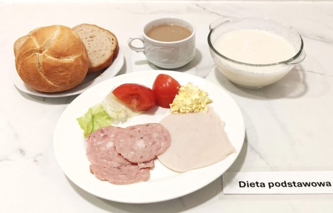 Talerz z plasterkami wędliny, sałatą, pomidorem i porcją masła. Obok kromka chleba, bułka kajzerka, filiżanka kawy z mlekiem oraz miseczka z białym napojem mlecznym. Na stoliku etykieta z napisem „Dieta podstawowa”