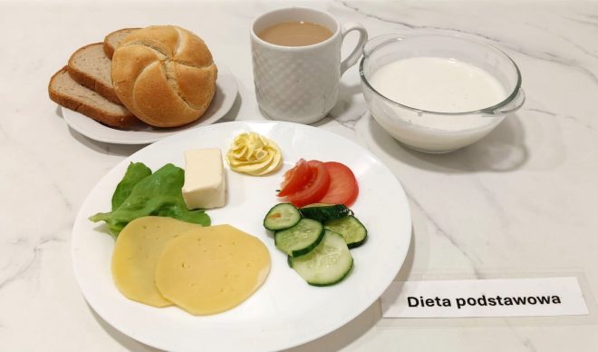 Śniadanie w diecie podstawowej: talerz z plastrami sera żółtego, sałatą, masłem, pomidorem, ogórkiem i porcją białego sera. Obok pieczywo: bułka i kromki chleba razowego. W tle filiżanka kawy z mlekiem oraz miseczka z białym jogurtem lub kefirem. Całość ułożona na marmurowym blacie, z etykietą „Dieta podstawowa”