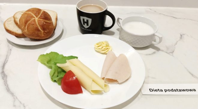Taca z podpisem „Dieta podstawowa”. Na talerzu znajdują się plasterki sera żółtego i wędliny drobiowej, kawałek pomidora, liść sałaty oraz porcja masła. Obok talerzyk z pieczywem: jedna kajzerka i dwie kromki chleba pszennego. W tle filiżanka z mlekiem i kubek z kawą z mlekiem lub herbatą mleczną