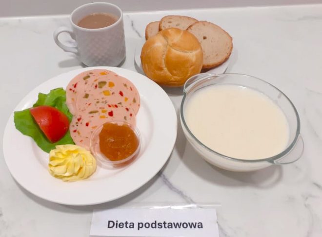 Talerz ze śniadaniem w diecie podstawowej: dwa plastry mortadeli z warzywami, sałata z kawałkiem pomidora, porcja masła i dżemu w pojemniku. Obok bułka, dwa kromki chleba, biały kubek z kawą z mlekiem oraz miseczka z białą zupą mleczną. Całość ustawiona na białym blacie. Etykieta z napisem „Dieta podstawowa”