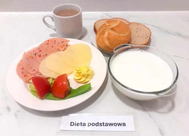 Talerz z plasterkami mortadeli z warzywami i sera żółtego, masło, sałata i pomidor. Obok bułka kajzerka i dwie kromki chleba pszennego. Filiżanka kawy z mlekiem i miseczka mleka. Dieta podstawowa