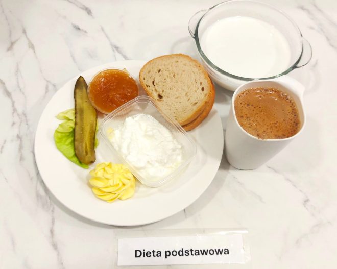 Zestaw śniadaniowy serwowany w ramach diety podstawowej. Na talerzu umieszczono trzy kromki chleba pszennego, porcję twarogu w plastikowym pojemniku, odrobinę masła w formie dekoracyjnej rozetki, plaster kiszonego ogórka na liściu sałaty oraz porcję marmolady lub dżemu o jasnym, morelowym kolorze. Do picia podano dwa napoje: kakao lub kawę zbożową z mlekiem – w białym kubku – oraz szklankę mleka w szklanym naczyniu z uchwytami. Całość zaprezentowana na marmurowym blacie z etykietą „Dieta podstawowa”
