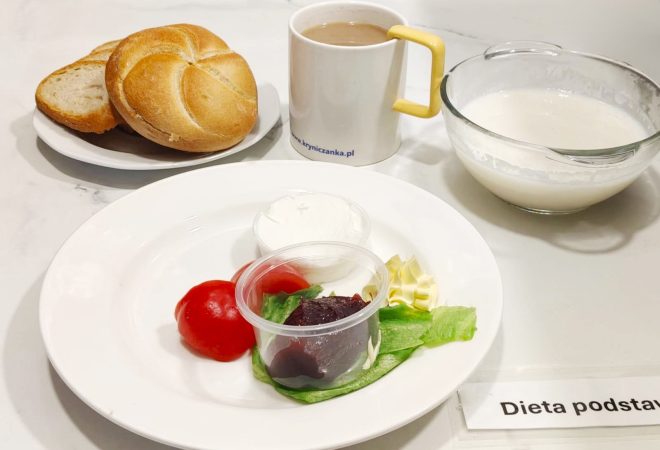 Talerz z pomidorem, liściem sałaty, porcją białego twarożku, masła oraz dżemu w plastikowym kubeczku. Obok bułka kajzerka i kromki chleba, kubek z kawą z mlekiem i miseczka z białym napojem mlecznym. Na stoliku etykieta z napisem „Dieta podstawowa”