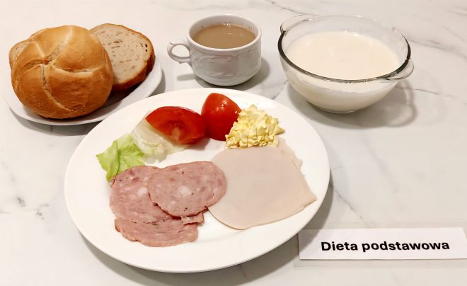 Na talerzu śniadaniowym ułożono dwa plasterki wędliny (jeden z mielonki, drugi z indyka), liść sałaty, dwie ćwiartki pomidora oraz porcję masła. W tle – bułka typu kajzerka i kromki chleba pszennego na osobnym talerzyku. Obok znajduje się filiżanka z kawą z mlekiem oraz szklana miska z porcją mleka. Przy zestawie leży etykieta „Dieta podstawowa”