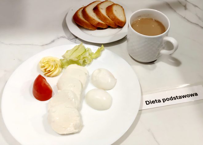 Zestaw śniadaniowy w diecie podstawowej. Na białym talerzu plasterki piersi z kurczaka w galarecie, dwa jajka ugotowane na twardo, liść sałaty, dekoracyjnie uformowane masło i kawałek pomidora. W tle osobny talerzyk z czterema kromkami chleba pszennego. Po prawej stronie filiżanka z kawą z mlekiem. Na blacie widoczna kartka z napisem „Dieta podstawowa”.
