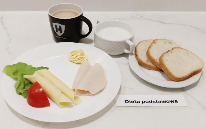 Zestaw śniadaniowy w diecie podstawowej. Na białym talerzu leżą dwa plastry sera żółtego, dwa plastry wędliny z kurczaka, kawałek pomidora i liść sałaty, a także porcja masła. Obok stoi kubek z kawą z mlekiem, miseczka z białym napojem (najpewniej mlekiem) oraz talerz z trzema kromkami jasnego pieczywa. Przed talerzem widoczna jest karteczka z napisem "Dieta podstawowa"