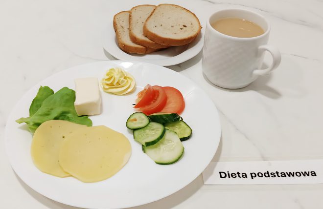 Białe porcelanowe talerze na jasnym blacie. Na głównym talerzu: dwa plasterki żółtego sera, liść sałaty, kostka białego sera, porcja masła w kształcie rozety, plasterki pomidora oraz kilka plasterków ogórka. Obok na osobnym talerzyku trzy kromki białego chleba. Do śniadania podano napój w białym kubku – najprawdopodobniej kawę z mlekiem. Na stoliku leży podpis: „Dieta podstawowa”